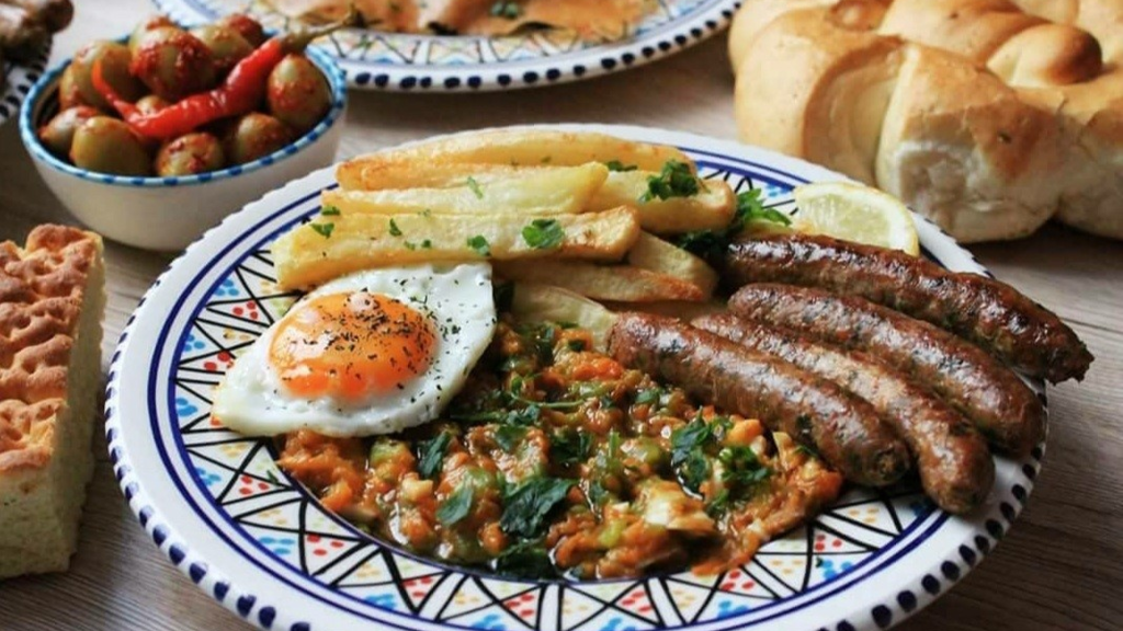 kafteji on plate with fried egg fries and sausages on table with other foods